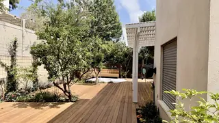 דק עץ בגינה לצד בית, עם פרגולה לבנה ברקע